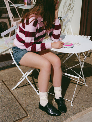 Teenage girl at a café table in a pink and burgundy striped jumper, denim shorts, cream socks and black soft crinkled patent Maévie Oxford Ballet flats