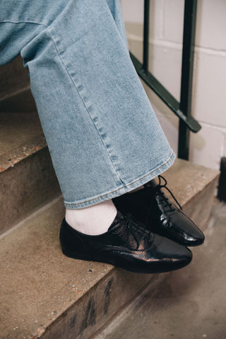 Black patent Maévie Oxford Ballet flats worn with white socks and light-wash jeans, resting on concrete steps